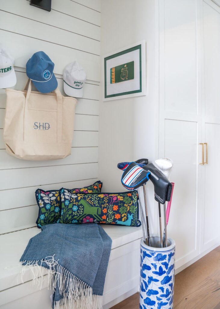 A mudroom corner with a white shiplap wall, tote bag, white hats, floral pillows on a bench, a blue throw, framed art, and a golf bag with clubs—styled by an interior designer Boerne TX—rests elegantly on the wooden floor.