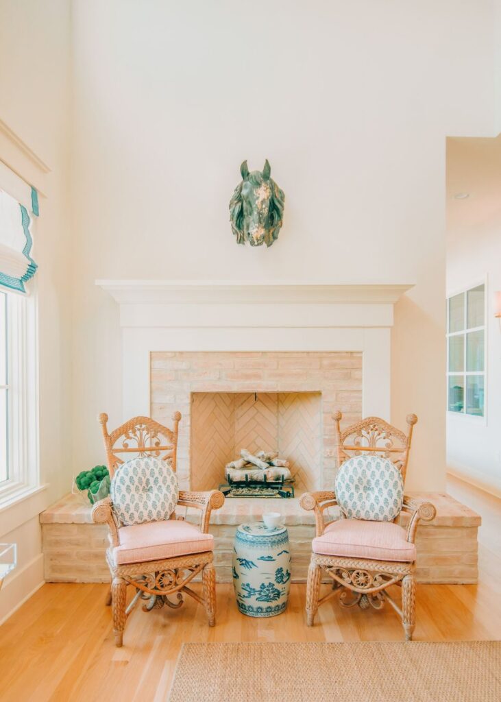 A bright living room styled by an interior designer Boerne TX, with two wicker chairs, patterned cushions, a blue-and-white ceramic stool, brick fireplace, greenery, and a decorative horse head above the mantel. Sunlight pours through the window.