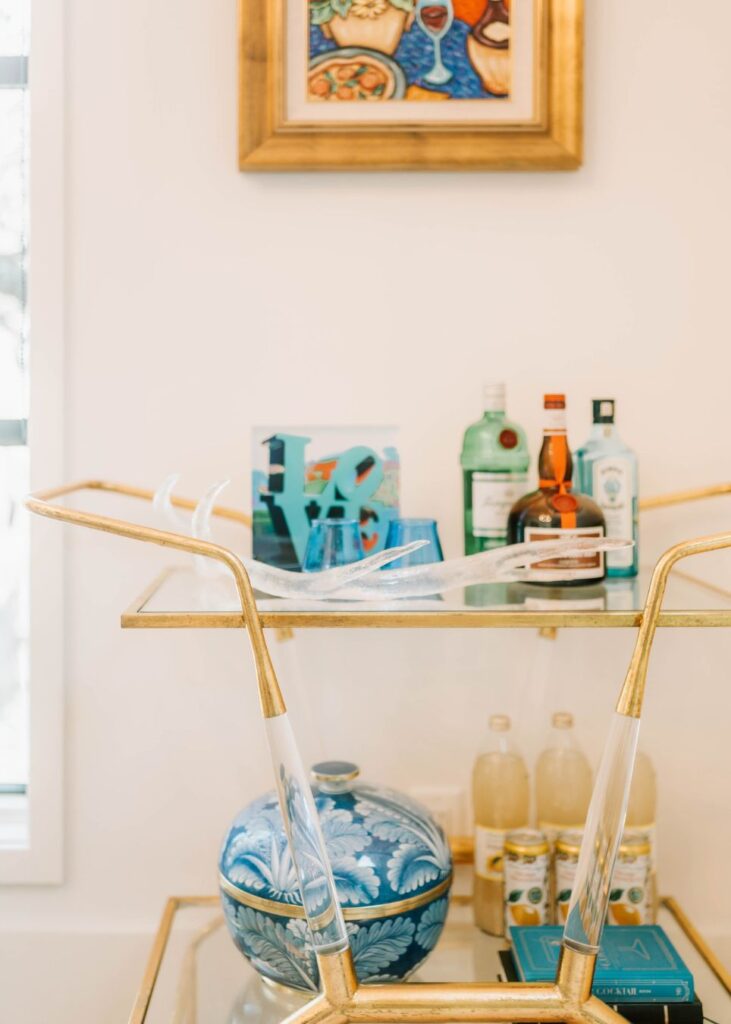 A gold and glass bar cart, styled by an interior design company in Boerne, TX, holds assorted liquor bottles, a blue 40 decoration, and glasses. The bottom shelf displays a blue patterned dish and lemonade bottles beneath colorful framed artwork.