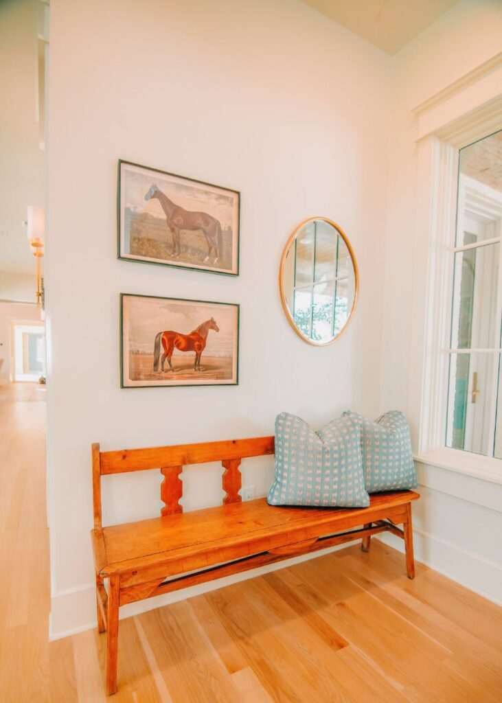 A wooden bench with two light blue patterned pillows sits against a white wall, styled by an interior designer in Boerne, TX. Two framed horse paintings and a round mirror decorate the space, as natural light streams in from a nearby window.