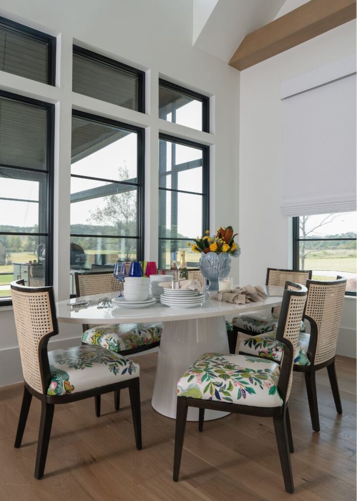 A modern dining area designed by a leading interior design company in Boerne, TX, features a round white table, colorful tableware, floral-patterned chairs, fresh flowers, large windows, and scenic views bathed in natural light.