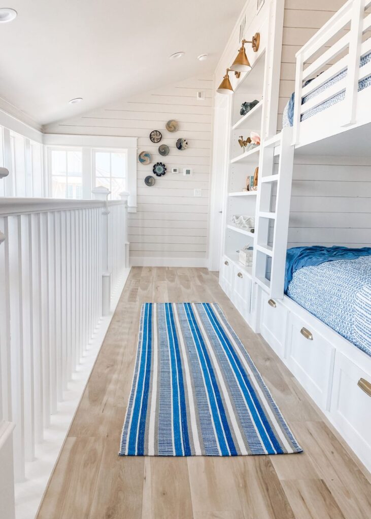 A bright, airy hallway by an interior design company in Boerne, TX, featuring wood floors, white built-in bunk beds with blue bedding, shiplap walls, a striped blue rug, shelves with decor, and nautical wall art above a row of windows.