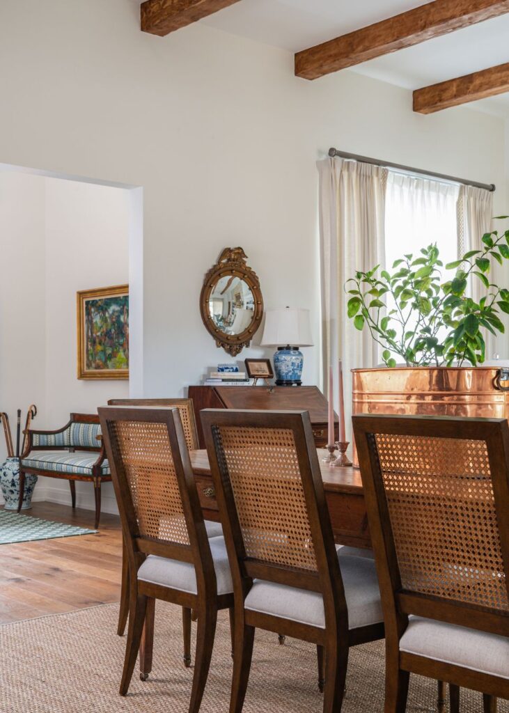 A bright dining room designed by an interior design company in Boerne, TX, featuring wooden beams, a wooden table and woven-back chairs, a large potted plant, a vintage mirror, and art on the walls. Sunlight streams in through a curtained window.