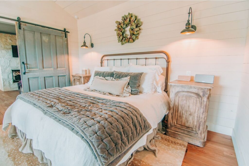 A cozy farmhouse-style bedroom designed by an interior decorator in Boerne, featuring a metal-frame bed, plush gray bedding, rustic wooden nightstands, wall-mounted lamps, a decorative wreath above the bed, and a sliding barn door.
