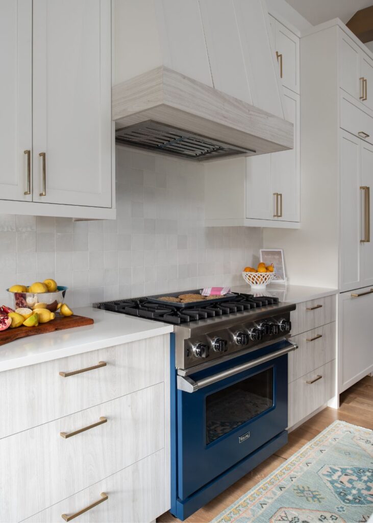 Modern kitchen with white cabinets, gold handles, a blue oven, marble-like countertops, and a white tile backsplash. Designed by an expert interior designer Boerne TX, it features fresh fruit on the counter and a blue patterned rug.