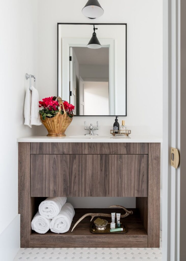 A modern bathroom with a wooden vanity, white countertop, and large mirror—styled by an interior designer Boerne TX. Accents include a basket of red flowers, toiletries, rolled white towels, and a tray with bathroom essentials.