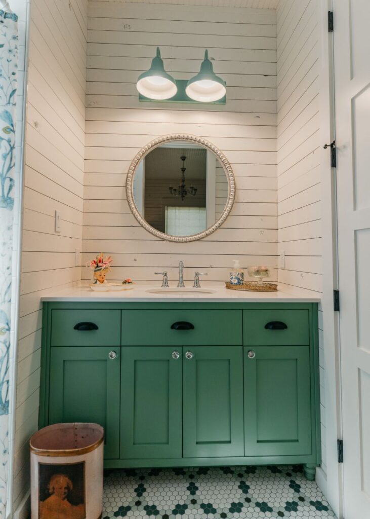 A small bathroom by an interior decorator Boerne features green cabinets, a round mirror with decorative frame, dual faucets, white shiplap walls, and green-and-white hexagon tile flooring illuminated by green-shaded lights.