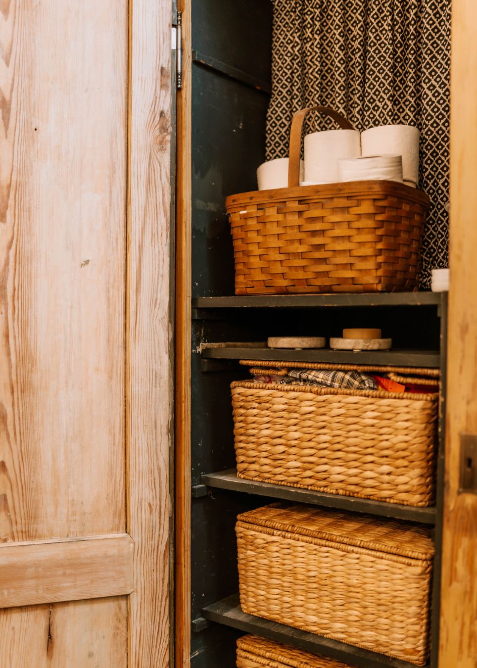 An open wooden cabinet with shelves holds woven baskets, rolled-up towels, and toilet paper rolls. Lined with a patterned fabric curtain, it reflects the thoughtful touch of an interior decorator Boerne would appreciate.