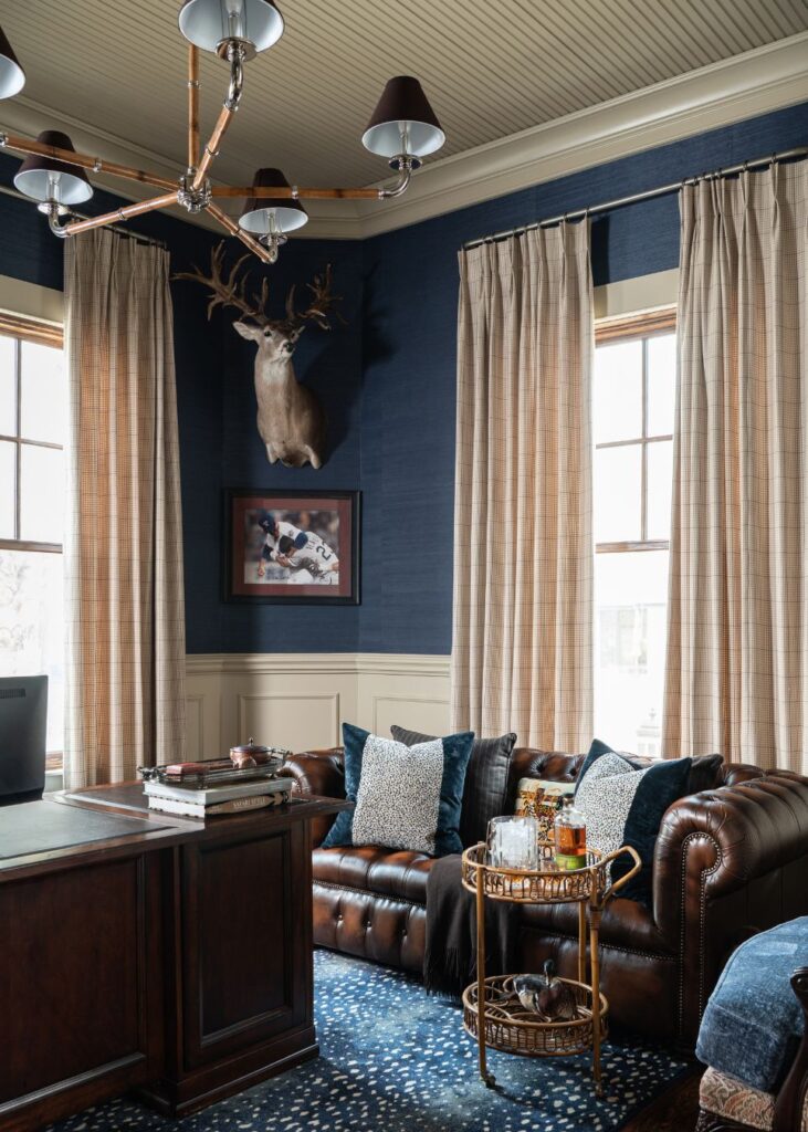 A stylish home office by an interior designer in Boerne, TX features a dark leather sofa with blue and white pillows, a wooden desk, tall windows with beige curtains, a bar cart, blue walls, a deer head mount, and a framed hockey photo.