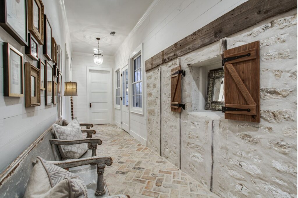 A rustic hallway designed by an interior decorator Boerne features a brick floor, white shiplap walls, stone accents, wooden ceiling beams, and wood-framed windows. A bench with pillows and a wall of framed pictures add cozy decor.