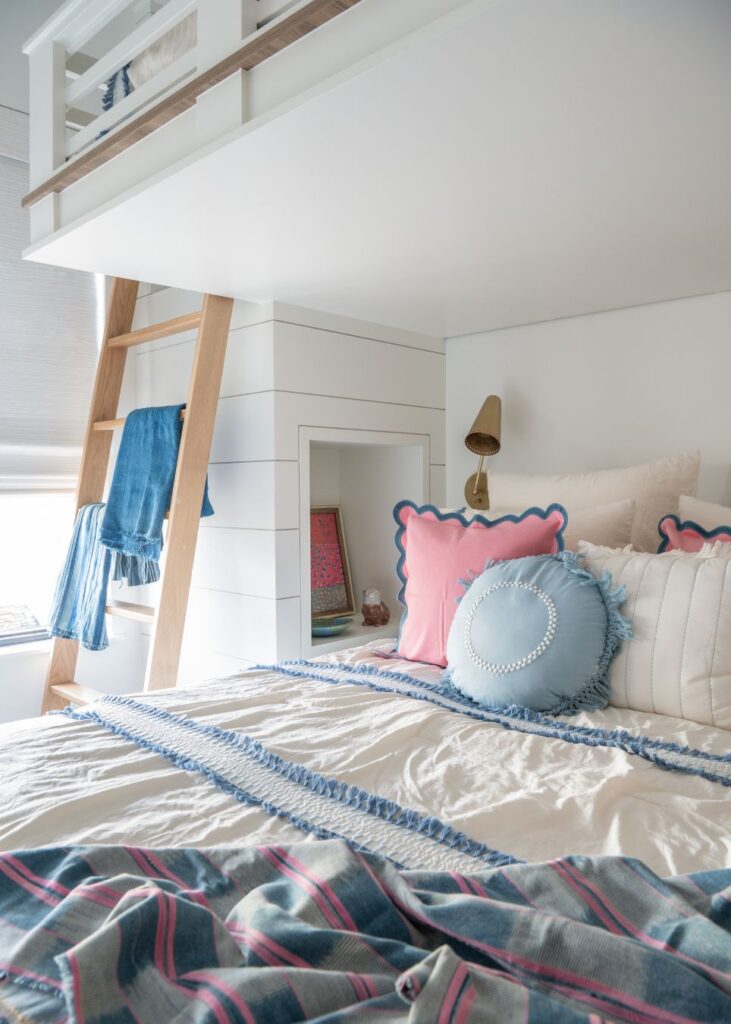 A cozy bedroom styled by an interior designer in Boerne, TX, featuring a loft bed ladder, soft pink and blue pillows, a tasseled blanket, plaid throw, and a built-in nook with books and a wall sconce.