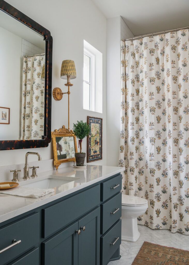 A bathroom with a dark blue vanity, marble countertop, and large mirror offers inspiring powder room design ideas. Decorative items, framed art, and a potted plant sit on the counter, while a white floral shower curtain and rug complete the cozy decor.