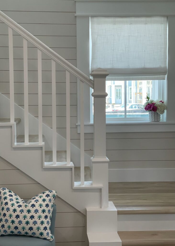 A white staircase with vertical railings stands beside a sunlit window draped in a sheer shade. A vase of pink and white flowers brightens the sill—an inviting touch crafted by an expert interior designer in Boerne, TX.