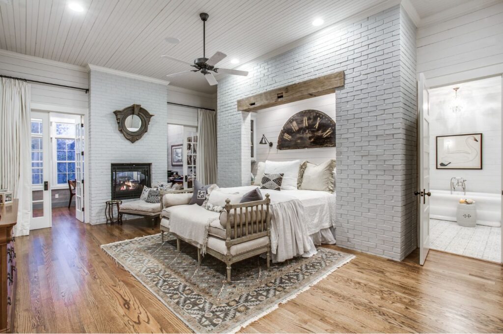 A cozy bedroom designed by an interior decorator Boerne features white brick walls, a wooden floor, a large bed with neutral bedding, a rug, sitting area, fireplace, French doors, and an adjoining bathroom with a freestanding tub.