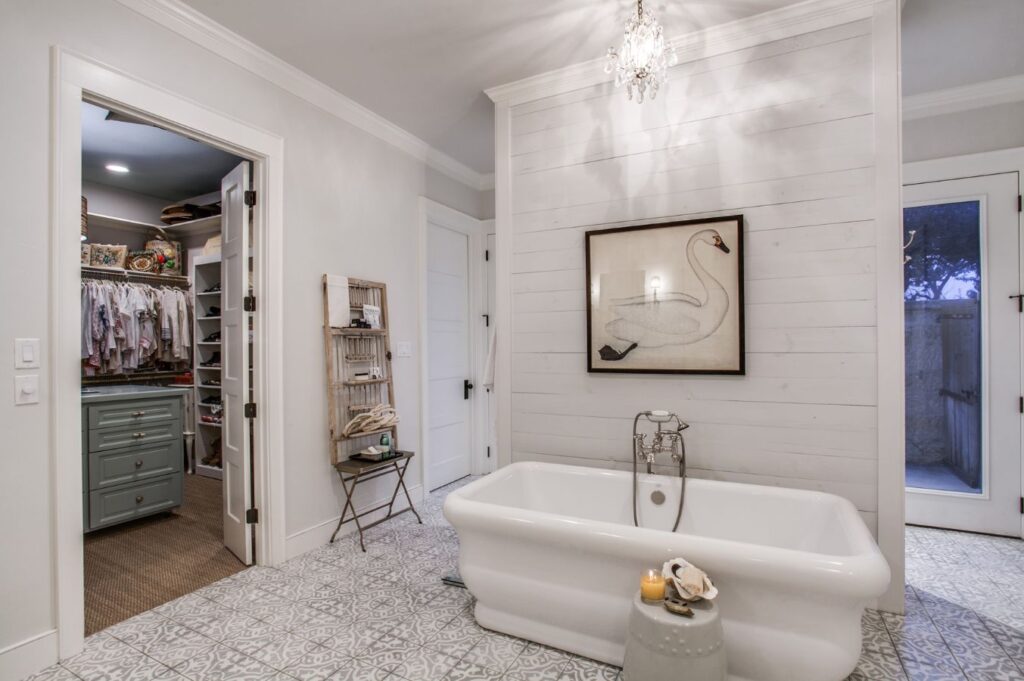 Elegant bathroom with a freestanding tub, patterned tile floor, chandelier, and swan artwork on a shiplap wall—crafted by an expert interior decorator Boerne. A closet with clothes is visible through an open door to the left.