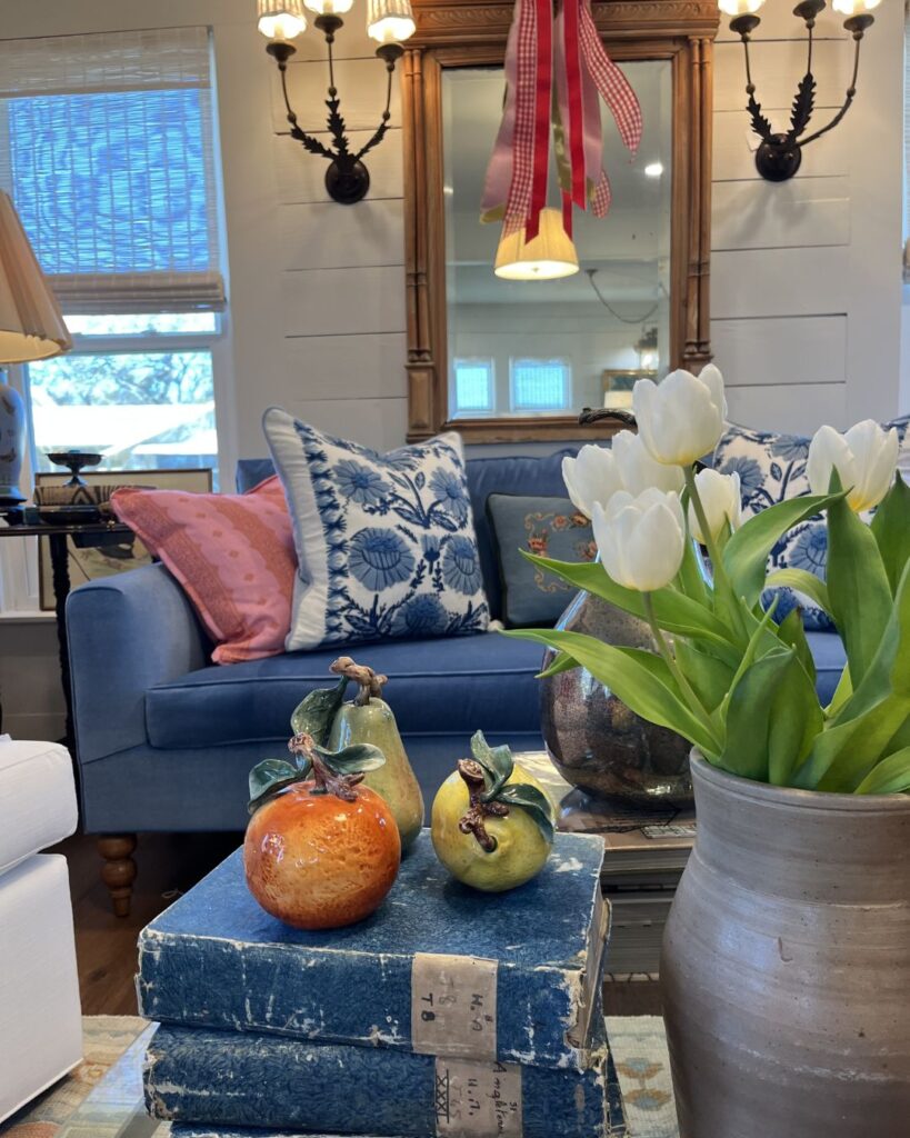 A cozy living room designed by an interior decorator in Boerne features a blue couch, patterned pillows, white tulips in a vase, ceramic fruit on vintage books, and a ribboned wall mirror lit by elegant sconces.