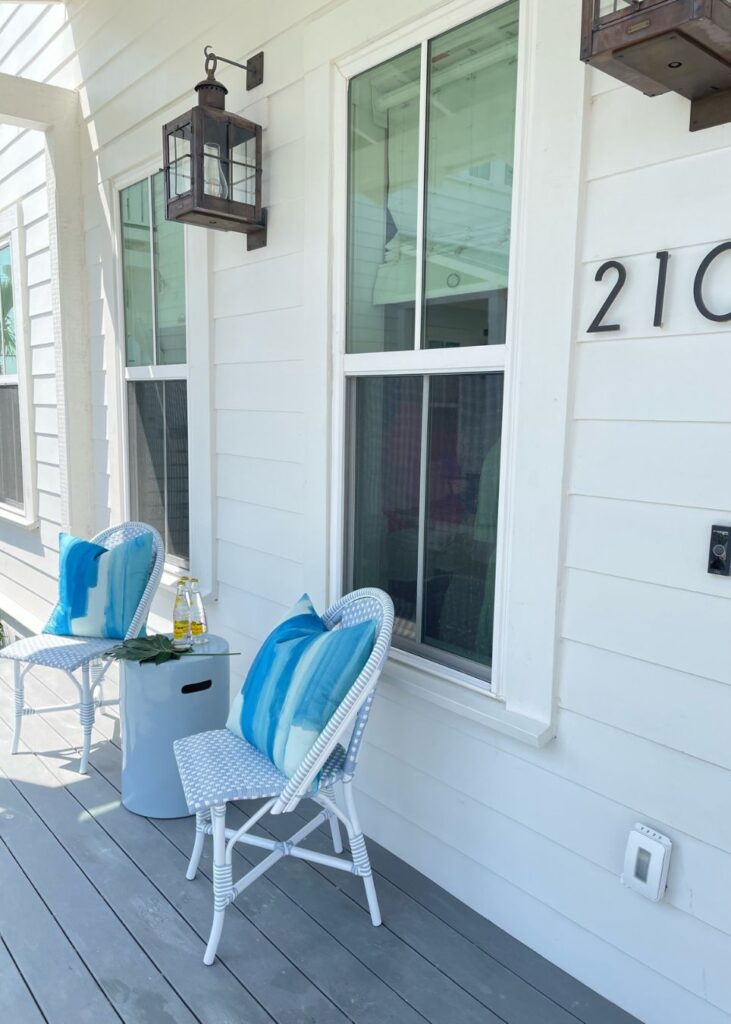 Two white wicker chairs with blue striped pillows sit on a porch beside a small white table holding lemonade and glasses. Styled by an interior designer Boerne TX, the home features white siding, large windows, and a charming lantern above the chairs.