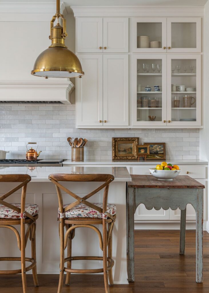 A bright farmhouse kitchen design with white cabinets, a subway tile backsplash, a brass pendant light, wooden stools with floral cushions, a rustic table, and a bowl of fruit on the counter. Framed art leans against the backsplash.