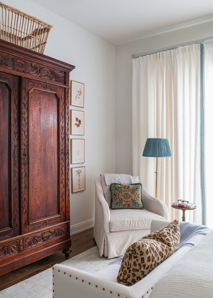 A cozy room styled by an interior designer Boerne TX, featuring a large wooden armoire, framed botanical prints, a white armchair with a floral pillow, a floor lamp with a blue shade, and a bed topped with a leopard print pillow.