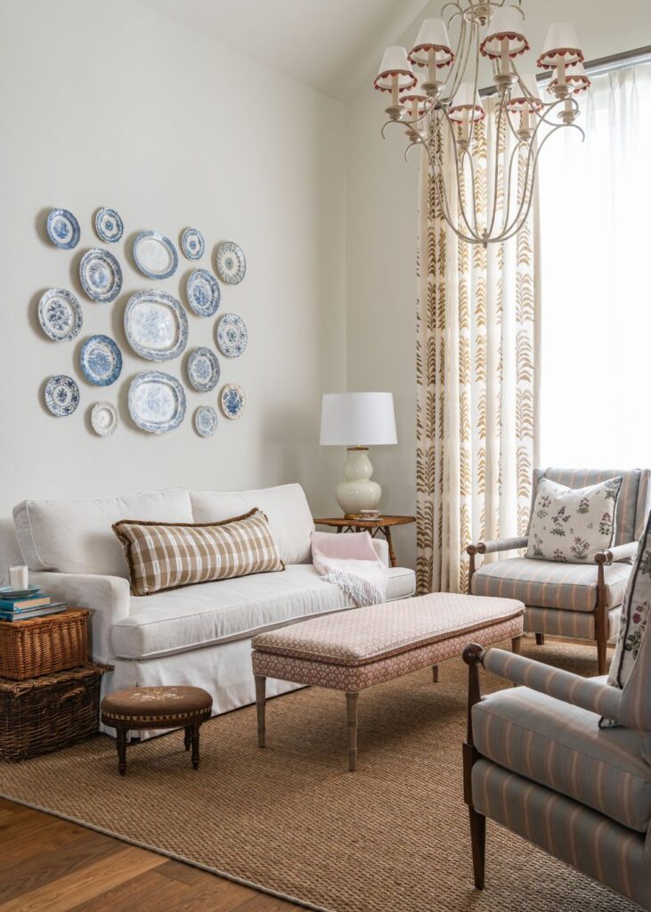 A cozy living room with a white sofa, striped pillows, patterned chairs, a woven rug, a bench coffee table, wall-mounted decorative plates, wicker accents, a table lamp, and a large chandelier near a window with patterned curtains.