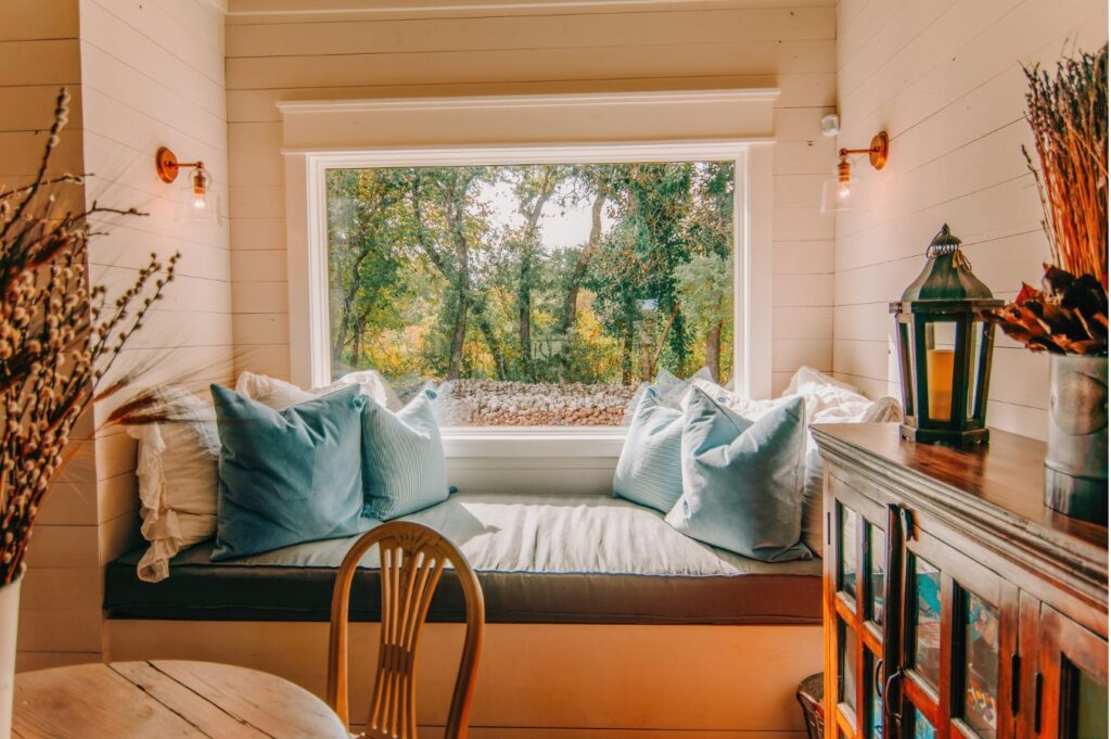 A cozy window nook with blue and white pillows overlooks a forest view—an inviting touch of interior design Boerne TX style, complete with warm lighting, a wooden chair, dried flowers, and a lantern on the cabinet.