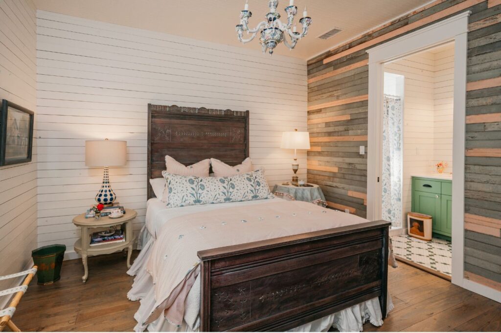 A cozy bedroom with white shiplap walls, a wooden bed with pastel bedding, elegant lighting, and a doorway to a bathroom with green cabinets—styled by an expert interior decorator Boerne for timeless comfort.