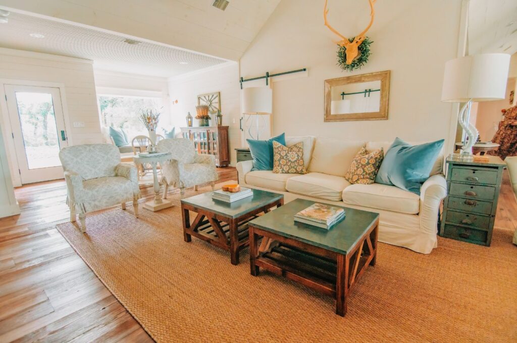 Bright, cozy living room with a white sofa, blue and patterned pillows, two armchairs, wooden coffee tables, woven rug, lamps, and wall decor. Sunlight streams in—styled by an expert interior designer Boerne TX—for a warm, inviting atmosphere.