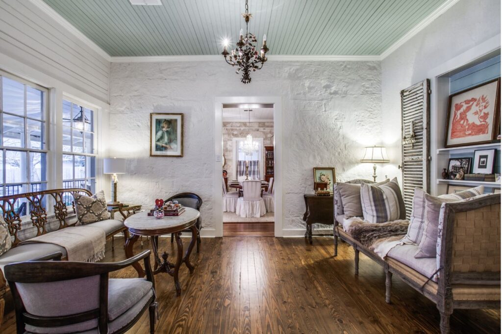A cozy vintage living room with wood floors, antique furniture, and a chandelier, beautifully styled by an interior designer Boerne TX. A doorway leads to a dining room with elegant touches, art, and lamps enhancing the inviting space.