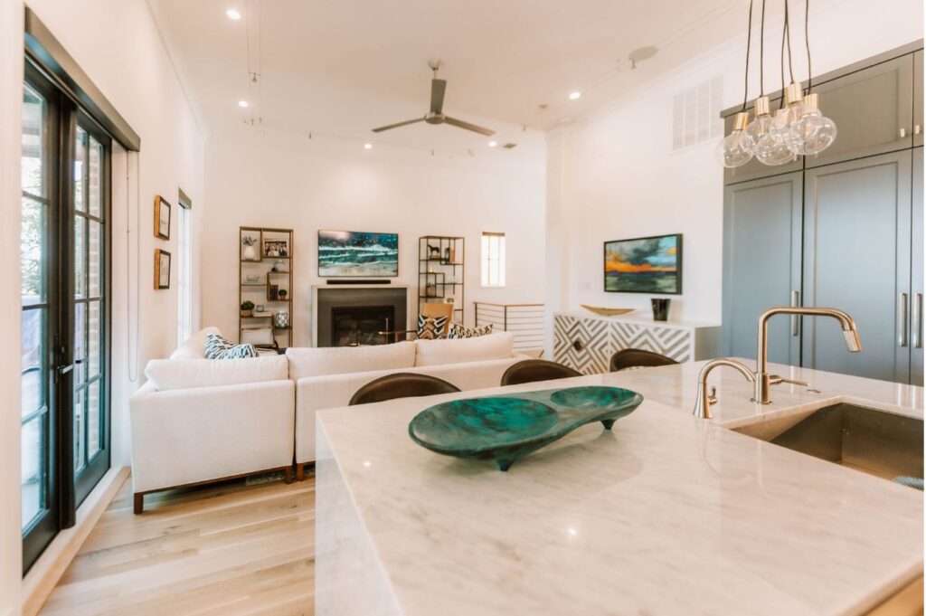 A modern living room designed by an interior decorator Boerne features light wood floors, a white sectional sofa, wall art, built-in shelves, a fireplace, flat-screen TV, and a marble kitchen island with a sink and decorative bowl in the foreground.