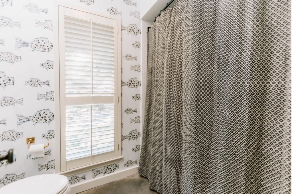 A bathroom designed by an interior design company in Boerne, TX, featuring patterned fish wallpaper, a window with white shutters, a toilet with toilet paper, and a black-and-white geometric shower curtain.