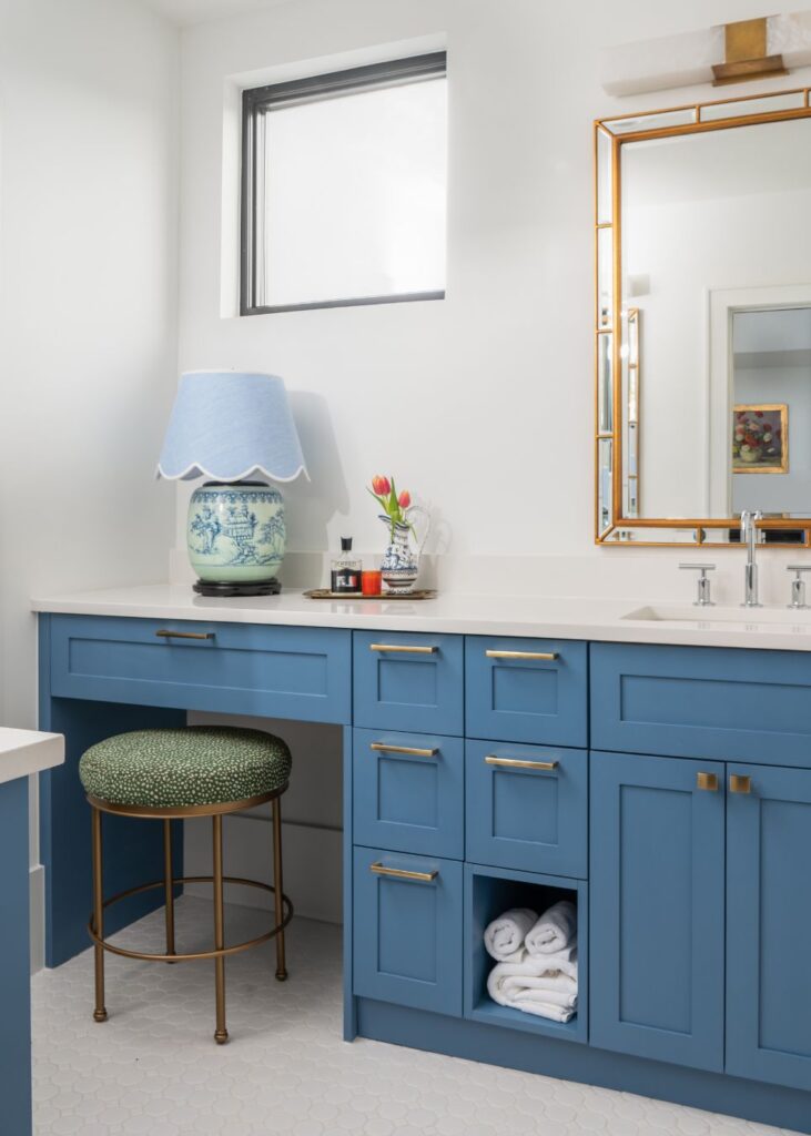 A bright bathroom by an interior designer Boerne TX features blue cabinets, a round green-cushioned stool, a countertop with a lamp, flowers, toiletries, a gold-framed mirror, rolled white towels, and a small window above.