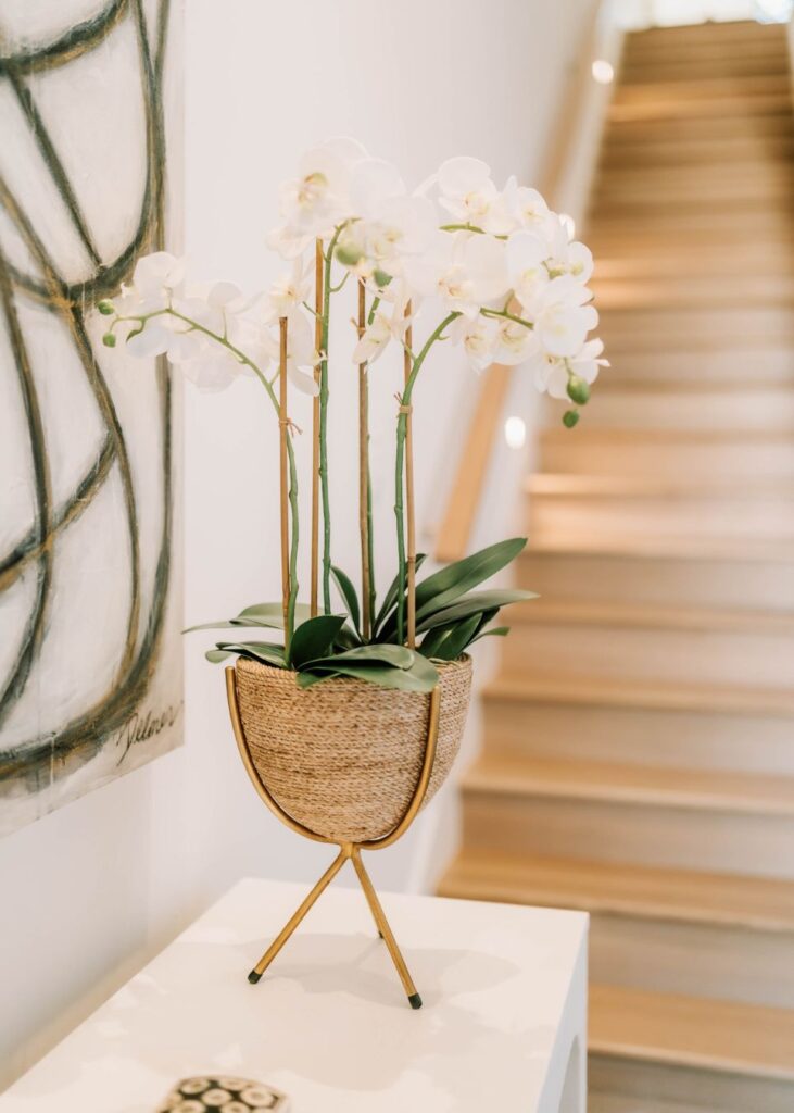 A potted white orchid with green leaves sits on a gold stand atop a white table. Behind it, a staircase with wooden steps leads upward—modern touches by an interior designer Boerne TX are evident, complemented by abstract art on the wall.