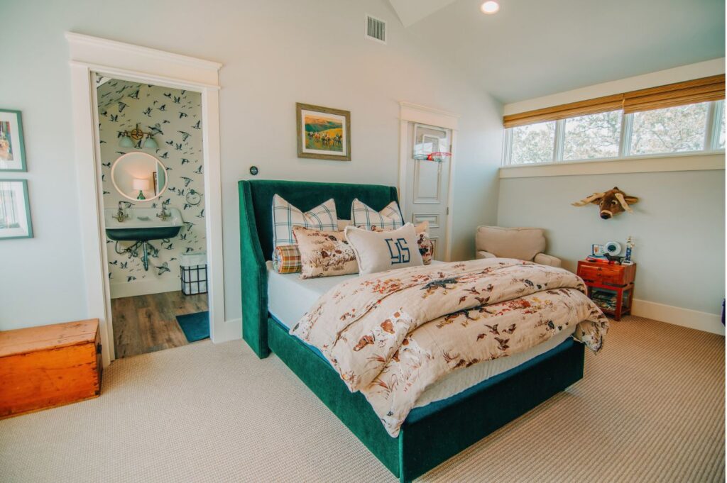 A cozy bedroom styled by an interior decorator in Boerne features a green upholstered bed, patterned bedding, plaid pillows, an armchair, wooden chest, mounted animal head, and art. An ensuite bathroom with bird wallpaper is visible.