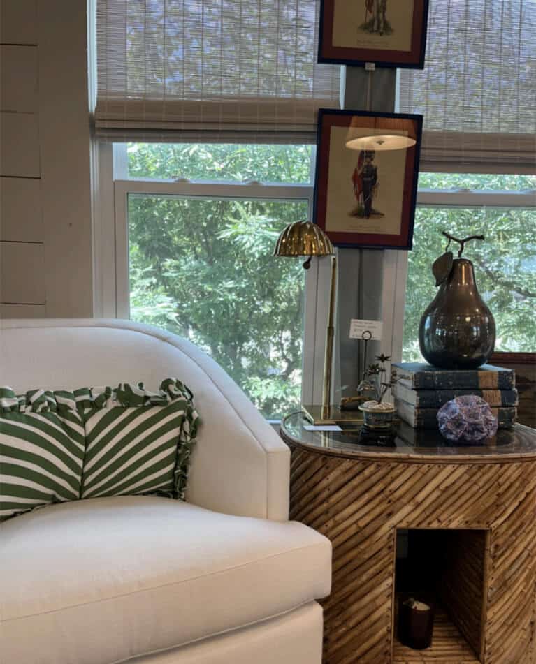 A white armchair with green and white striped pillows sits beside a round wooden side table holding books, a lamp, and decor. Large windows with blinds reveal leafy trees—a cozy look by an interior designer Boerne TX loves.