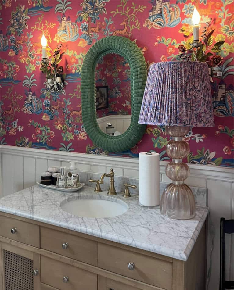 A bathroom vanity by an interior designer Boerne TX features a marble countertop, gold faucet, paper towel roll, green-framed oval mirror, floral sconces, and a pleated purple lamp set against pink patterned wallpaper.