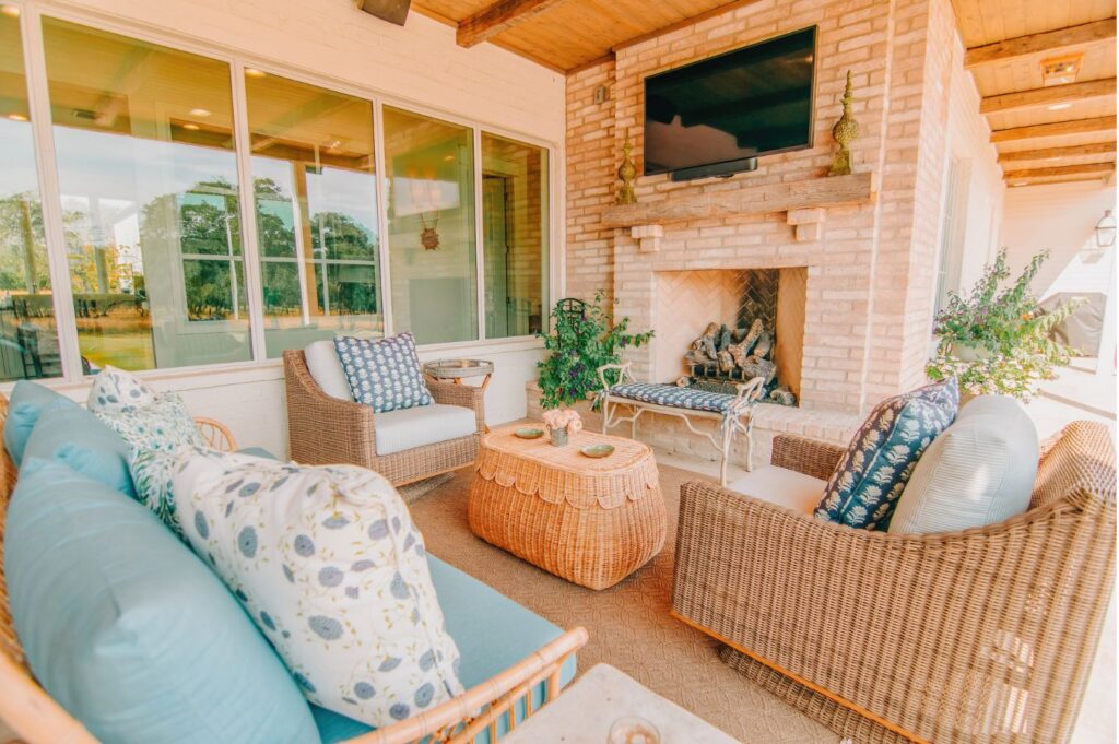 A cozy outdoor patio with wicker furniture, patterned cushions, and a brick fireplace designed by an interior design company Boerne TX. Large windows reflect lush greenery as soft natural light fills the inviting space.
