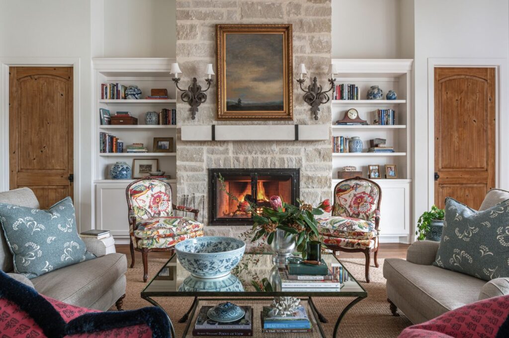 A cozy living room designed by an interior decorator in Boerne, featuring a stone fireplace, a painting above the mantel, floral armchairs, a glass coffee table with books and a blue bowl, built-in shelves, and wooden doors on either side.