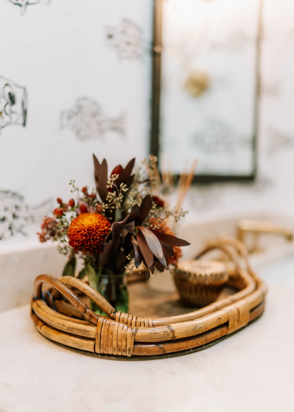 A rattan tray on a bathroom counter holds a small vase with dark red and orange flowers and a decorative container. Styled by an interior decorator in Boerne, the background features a black-and-white fish pattern and a blurred mirror.