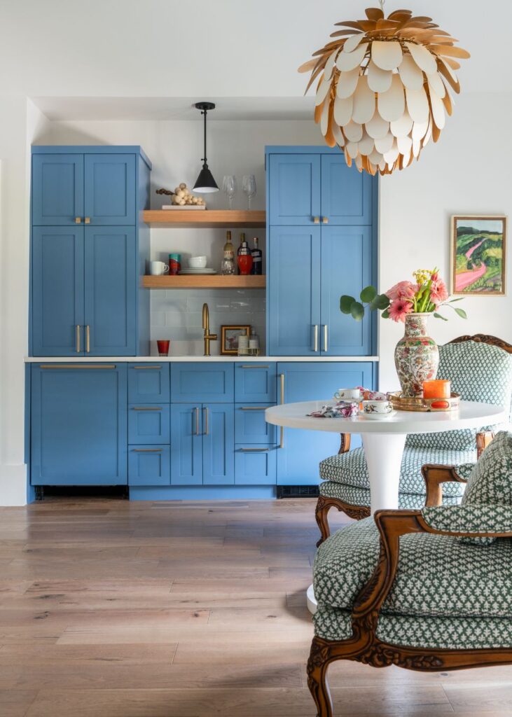 A stylish kitchen designed by an interior design company in Boerne, TX, features blue cabinets, open wood shelves with decor, a gold faucet, and a white round table with patterned armchairs under a modern flower-shaped light fixture.