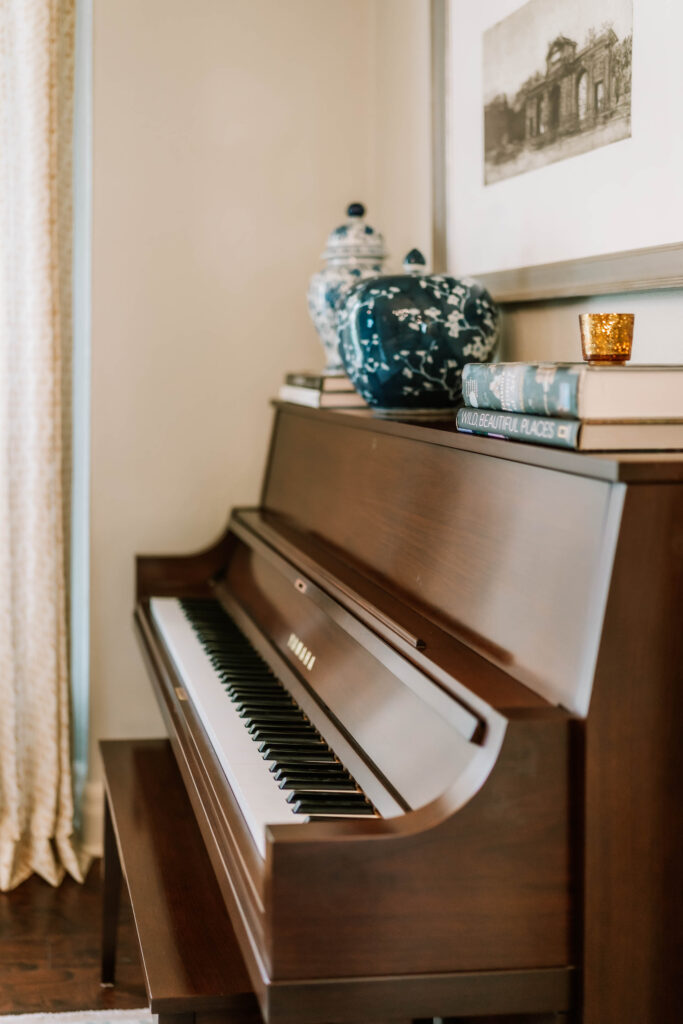 A brown upright piano sits against a wall, topped with stacked books, two blue and white vases, and a gold candle. A framed black-and-white art print hangs above—a cozy scene created by a skilled interior decorator Boerne. Light filters through the nearby curtain.