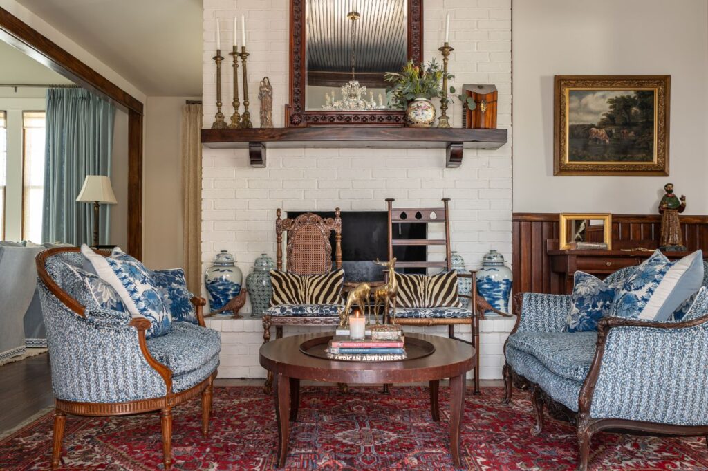 A traditional living room designed by an interior designer Boerne TX, featuring patterned blue armchairs, a wooden coffee table with books, a red rug, white brick fireplace, large mirror, decorative vases, and art on the walls.