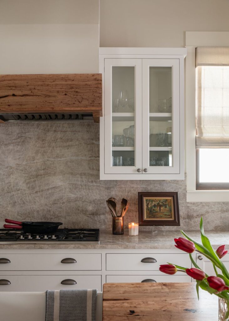A modern kitchen with white cabinets, a wood-accented range hood, and beige stone backsplash. Styled by an interior designer Boerne TX, it features red tulips, framed art, and natural light from a window with a fabric shade.