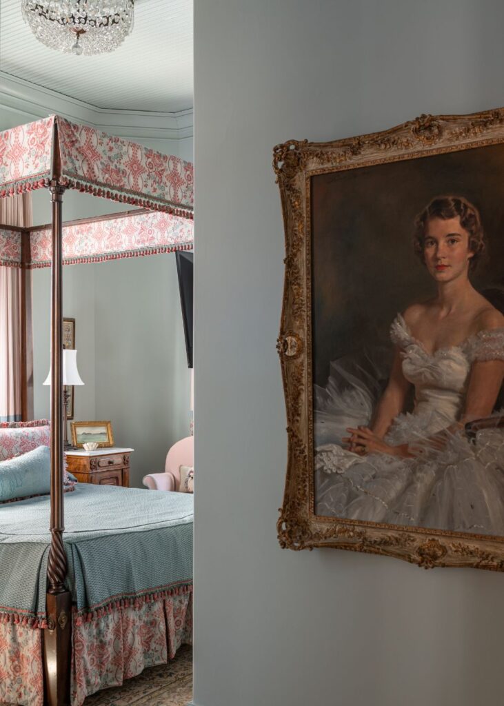 A framed portrait of a woman in a white dress hangs on a pale blue wall beside a traditional bedroom, elegantly styled by an interior designer Boerne TX, featuring a four-poster canopy bed, blue bedding, and patterned drapes.