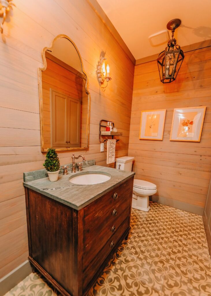 A cozy bathroom with wood-paneled walls, patterned tile floor, a dark wood vanity with a green countertop, gold-framed mirror, wall-mounted lights, and framed art above the towel rack—perfect inspiration from an interior decorator Boerne homeowners trust.