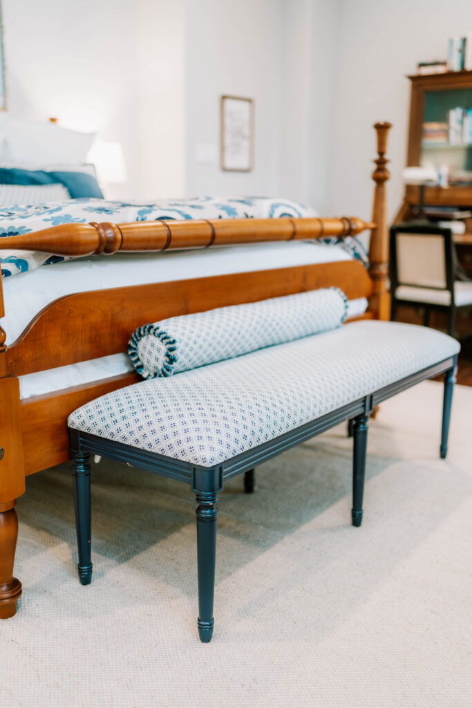 A wooden bed with blue and white bedding sits beside a patterned bench with cylindrical pillows in a cozy, well-lit bedroom—showcasing the touch of an interior designer Boerne TX, with a stylish desk in the background.