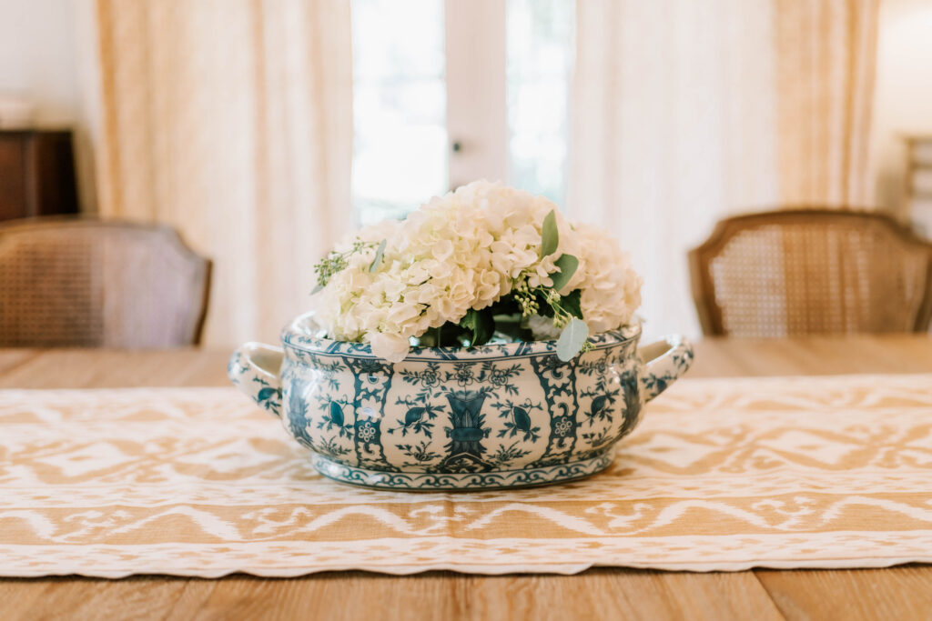 A blue and white ceramic vase with floral patterns holds white hydrangeas, sitting on a wooden dining table styled by an interior designer Boerne TX. Wicker chairs and cream curtains complete the elegant look.