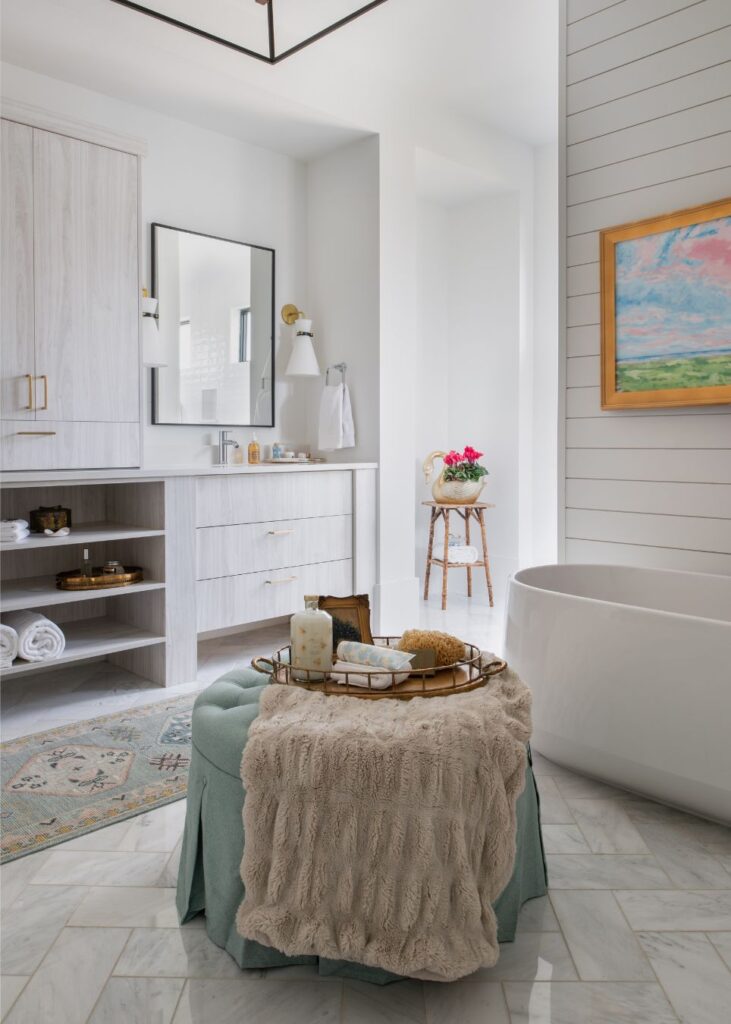 A modern bathroom designed by an interior decorator in Boerne features white cabinets, a round bathtub, a large mirror, and artwork on the shiplap wall. A tray with towels sits on a soft ottoman beside a small table with flowers.