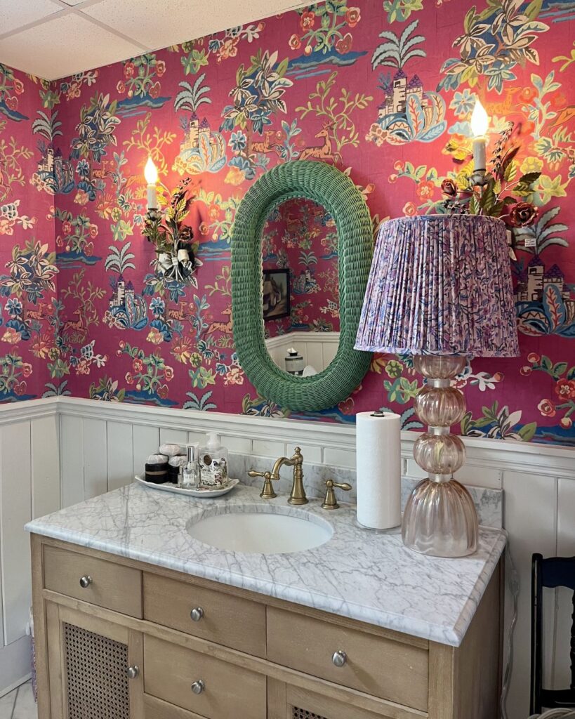 A bathroom with floral pink wallpaper, a marble countertop vanity, green oval mirror, gold faucet, and white wainscoting reflects the charm that an interior decorator Boerne can bring. Two wall sconces frame the mirror above a glass lamp with a purple patterned shade.