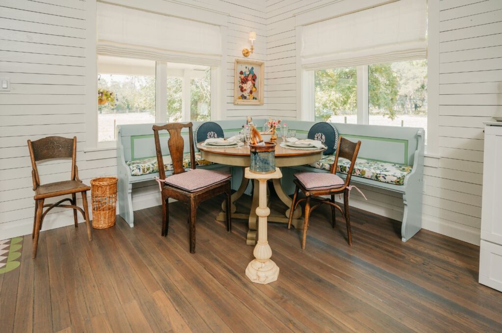 A cozy breakfast nook by an interior designer Boerne TX, with a round wooden table, mixed chairs, and a built-in green bench with floral cushions. Sunlight pours through large windows over white shiplap walls and hardwood floors.
