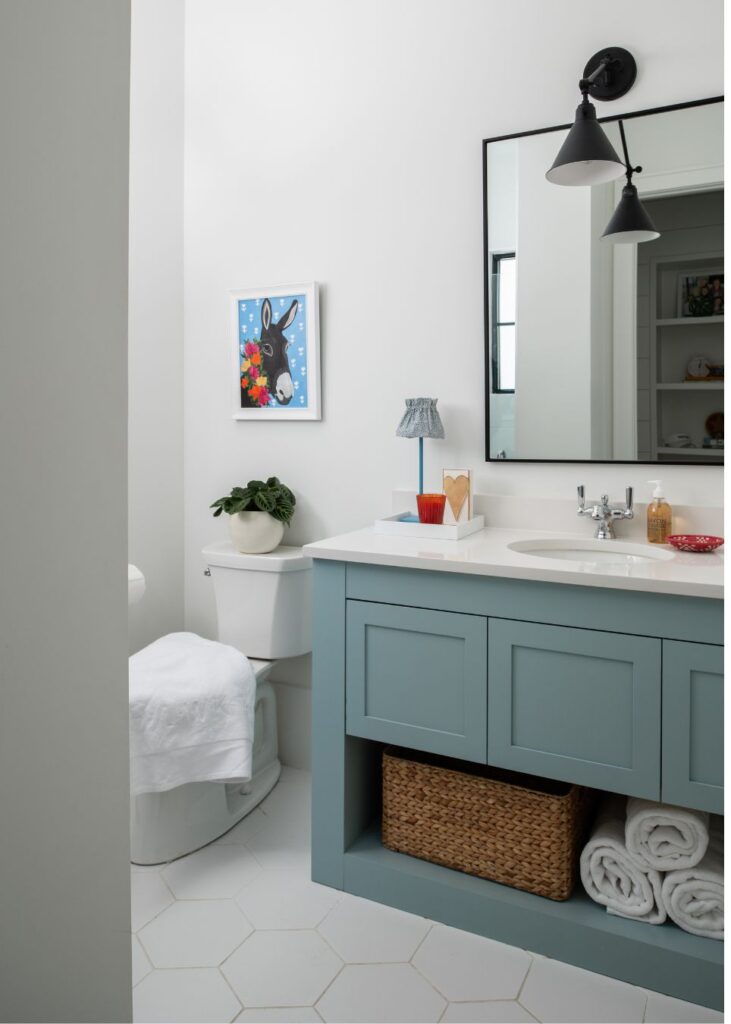A modern bathroom by an interior design company in Boerne, TX features a pale blue vanity, large mirror, and black sconce lighting. Towels and a wicker basket sit below the sink, with a plant on the toilet tank and a rabbit painting on the wall.